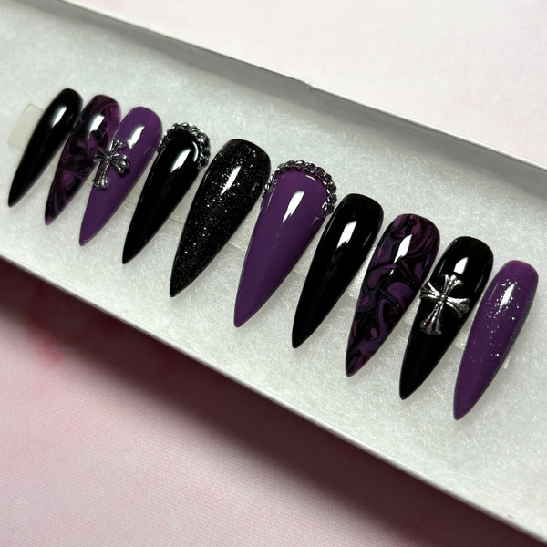 Load image into Gallery viewer, A set of handmade false nails in a long stiletto shape, featuring a gothic design in black and deep purple with a black-purple marble effect, subtle black and purple glitter, and 3D silver chain and cross embellishments, displayed in a white presentation box.
