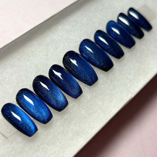 Load image into Gallery viewer, Blue cat-eye press-on nails that colour-shift from a shimmery dark blue through black, handmade in a short coffin shape, displayed in a white presentation box.
