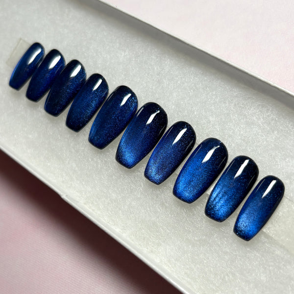 Load image into Gallery viewer, A set of blue cat-eye handmade press-on nails that colour-shift from a shimmery dark blue through black, in a short coffin shape, displayed in a white presentation box.
