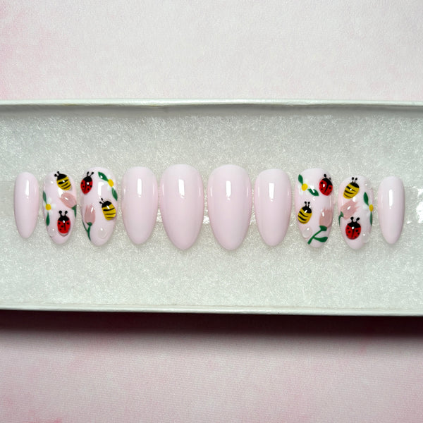 Load image into Gallery viewer, Pastel pink press-on nails featuring 3D bees, ladybirds and flowers on four of the nails, in a short almond shape, displayed in a white presentation box on a pink background.
