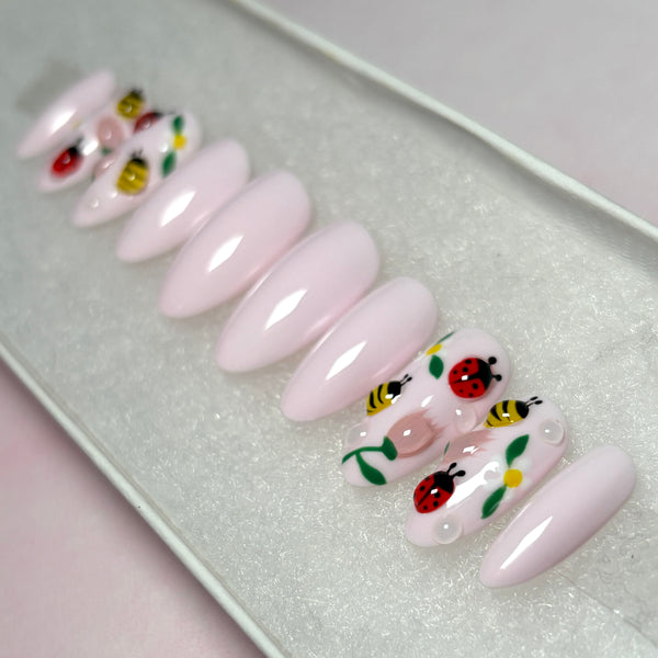 Load image into Gallery viewer, Soft pink press-on nails with accent nails featuring 3D ladybirds, bees and flowers artwork, in a short almond shape, displayed in a white presentation box.
