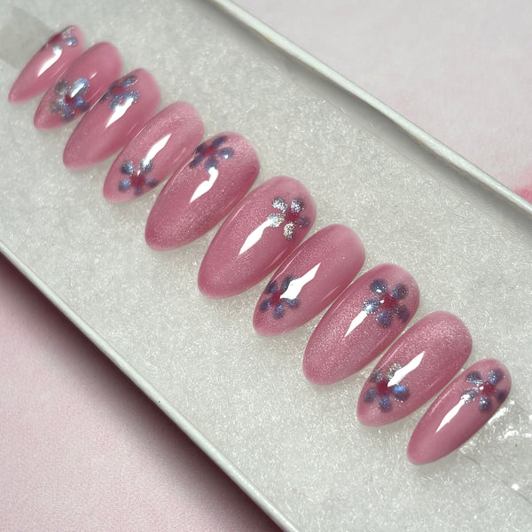 Load image into Gallery viewer, Short almond spring-summer press-on nails in a shimmery blush pink, with a lilac-silver flower on each nail in cat eye polish, displayed in a white presentation box.
