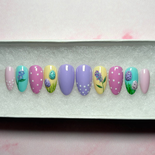 Load image into Gallery viewer, Easter press-on nails in a short almond shape, in soft pastel shades of lilac, pink, blue and yellow, featuring tiny 3D Easter eggs in green grass, a delicate floral design, and white polka dots on some nails, from Illuminate by Liana.
