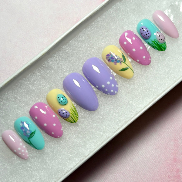 Load image into Gallery viewer, Short almond Easter press-on nails with 3D mini speckled eggs in green grass, a delicate floral design on two nails, and white polka dots on some nails, in pastel shades of lilac, pink, blue and yellow, in a white presentation box.

