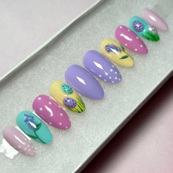 Load image into Gallery viewer, Easter press-on nails in pastel shades of lilac, pink, blue and yellow with 3D mini Easter eggs in grass, a delicate floral design, and white polka dots on some nails, in a short almond shape.

