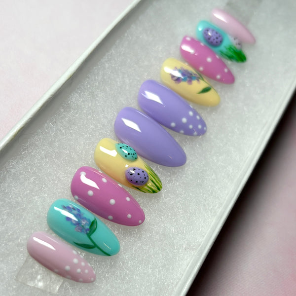 Load image into Gallery viewer, Easter press-on nails in pastel shades of lilac, pink, blue and yellow with 3D mini Easter eggs in grass, a delicate floral design, and white polka dots on some nails, in a short almond shape.
