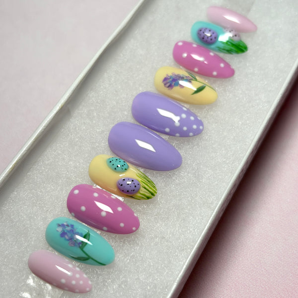 Load image into Gallery viewer, Handmade Easter fake nails with 3D mini Easter eggs in grass, a delicate floral design, and white polka dots on some nails, in pastel shades of lilac, pink, blue and yellow, in a short almond shape.

