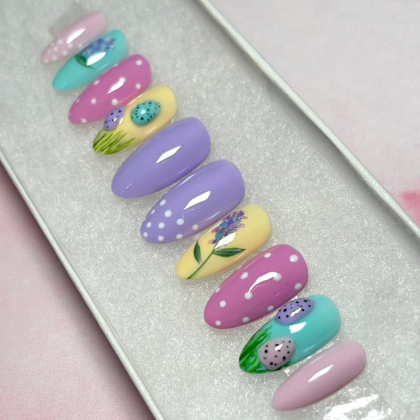 Load image into Gallery viewer, Handmade Easter press-on nails featuring 3D mini Easter eggs in green grass, a delicate floral design, and white polka dots on some nails, in pastel shades of lilac, pink, blue and yellow.
