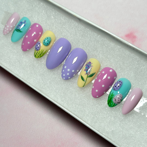 Load image into Gallery viewer, Easter press-on nails with 3D mini Easter eggs in grass, a delicate floral design, and white polka dots on some nails, in pastel shades of lilac, pink, blue and yellow, in a short almond shape.
