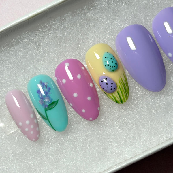 Load image into Gallery viewer, A close-up of handmade Easter press-on nails featuring two 3D mini Easter eggs in green grass on a pastel yellow nail, a delicate floral lavender design on a blue nail, and lilac and pink nails with polka dots, created by Illuminate by Liana UK. 
