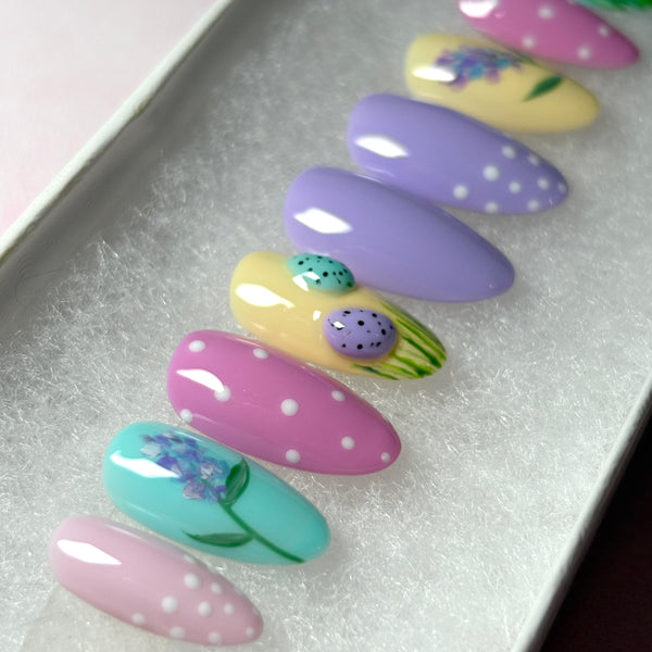 Load image into Gallery viewer, A close-up of Easter press-on nails with 3D mini Easter eggs in green grass on a pastel yellow nail, a delicate floral design on a blue nail, and white polka dots on pink and lilac, in a short almond shape. 

