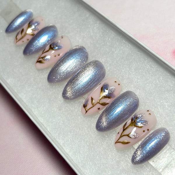 Load image into Gallery viewer, Short almond press-on nails with a delicate floral design on four nails featuring gold chrome stems and leaves and shimmering lilac petals on a soft pink base, with some nails in lilac cat-eye polish.
